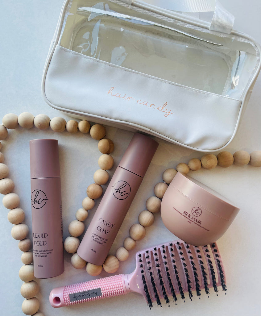 A collection of hair care products including two bottles of hair mask and oil, a brush, and a toiletry bag with handles, all displayed on a white surface with beads around.
